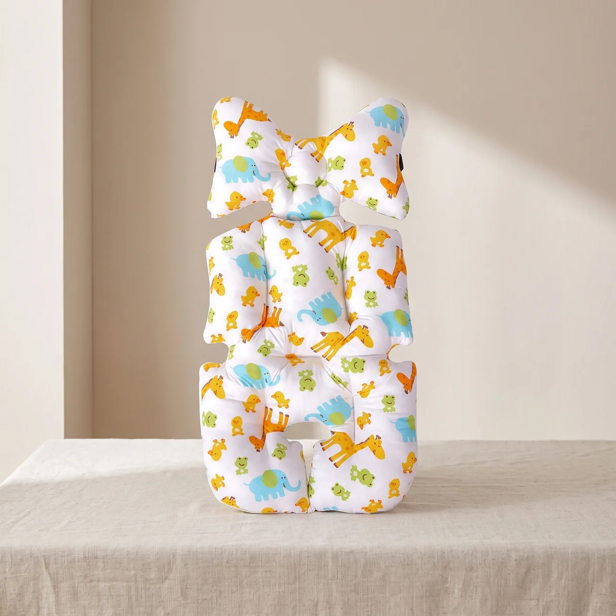 A baby support pillow with a dinosaur and elephant pattern, styled in a stacked position on a beige fabric-covered table against a neutral beige wall.