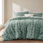 A sage green textured comforter set with matching pillow shams and a small white accent pillow, styled on a bed in a softly lit bedroom.
