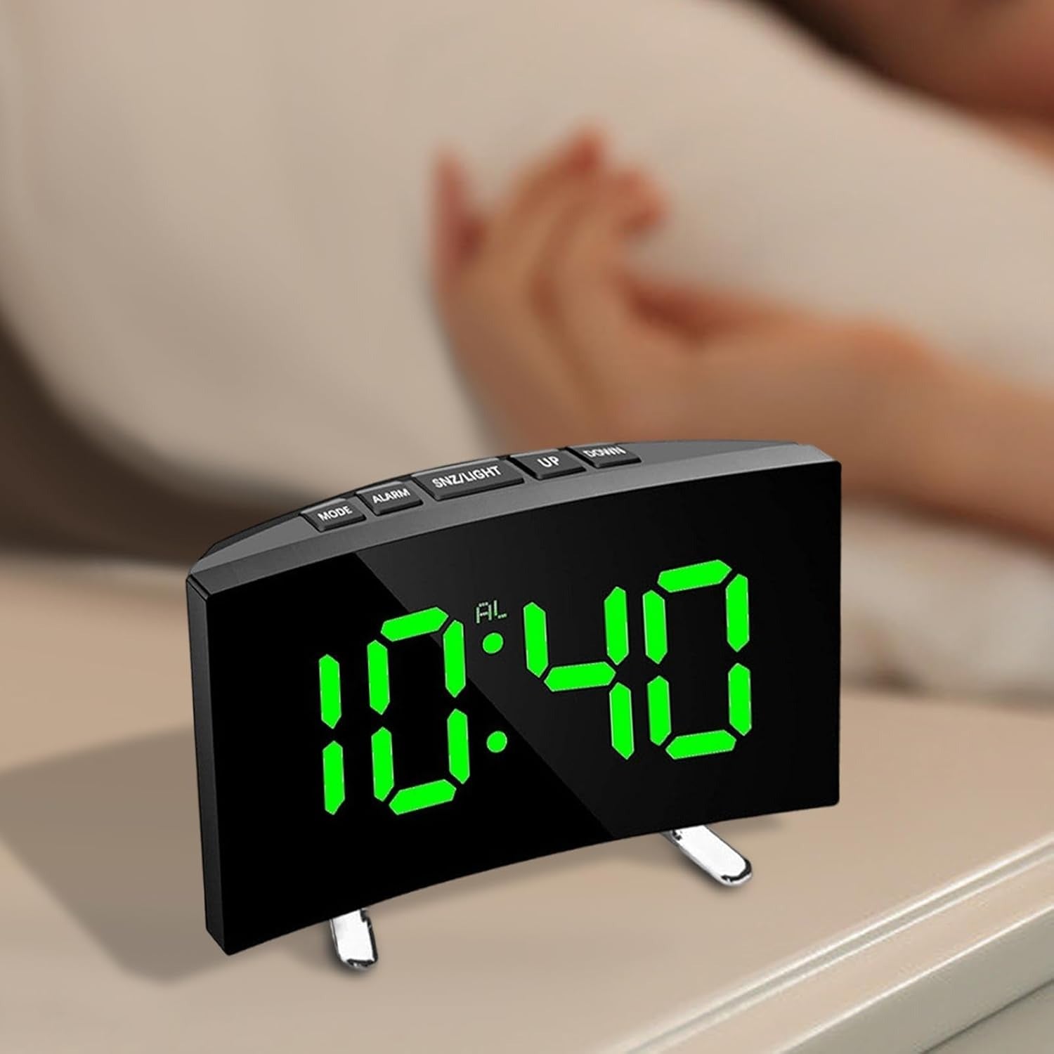 Digital clock with green LED display on a surface, blurred background of a person lying down.