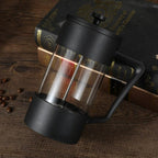 Black French press coffee maker on a wooden surface with coffee beans and books in the background
