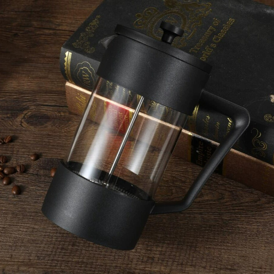 Black French press coffee maker on a wooden surface with coffee beans and books in the background