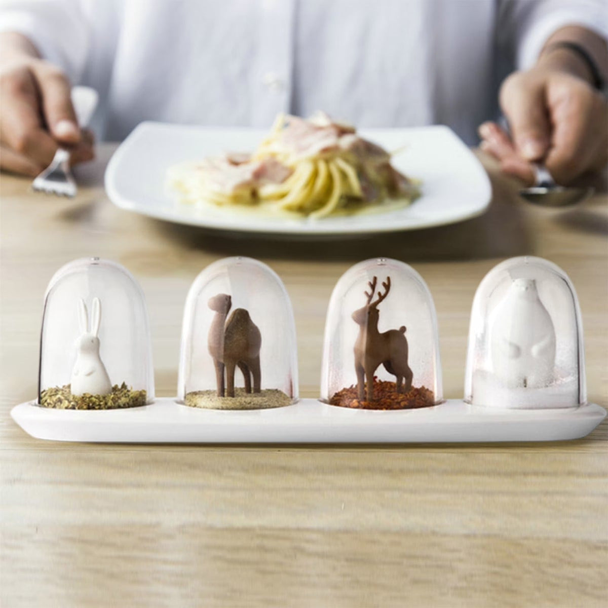 Four glass dome ornaments with miniature animal figurines on a white tray, with pasta, plate, fork, and spoon in the background.