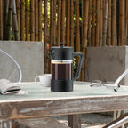 French press with coffee on a table outdoors, surrounded by bamboo and a chair.