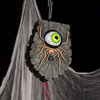 A Halloween hanging decoration shaped like a stone wall with a large realistic spider and glowing green eye motif, suspended against a black background with cobweb props.