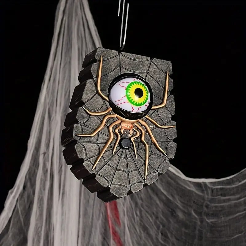 A Halloween hanging decoration shaped like a stone wall with a large realistic spider and glowing green eye motif, suspended against a black background with cobweb props.