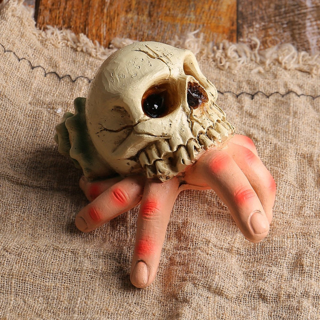 A Halloween prop of a human hand gripping a realistic skull, resting on a textured beige burlap fabric background.