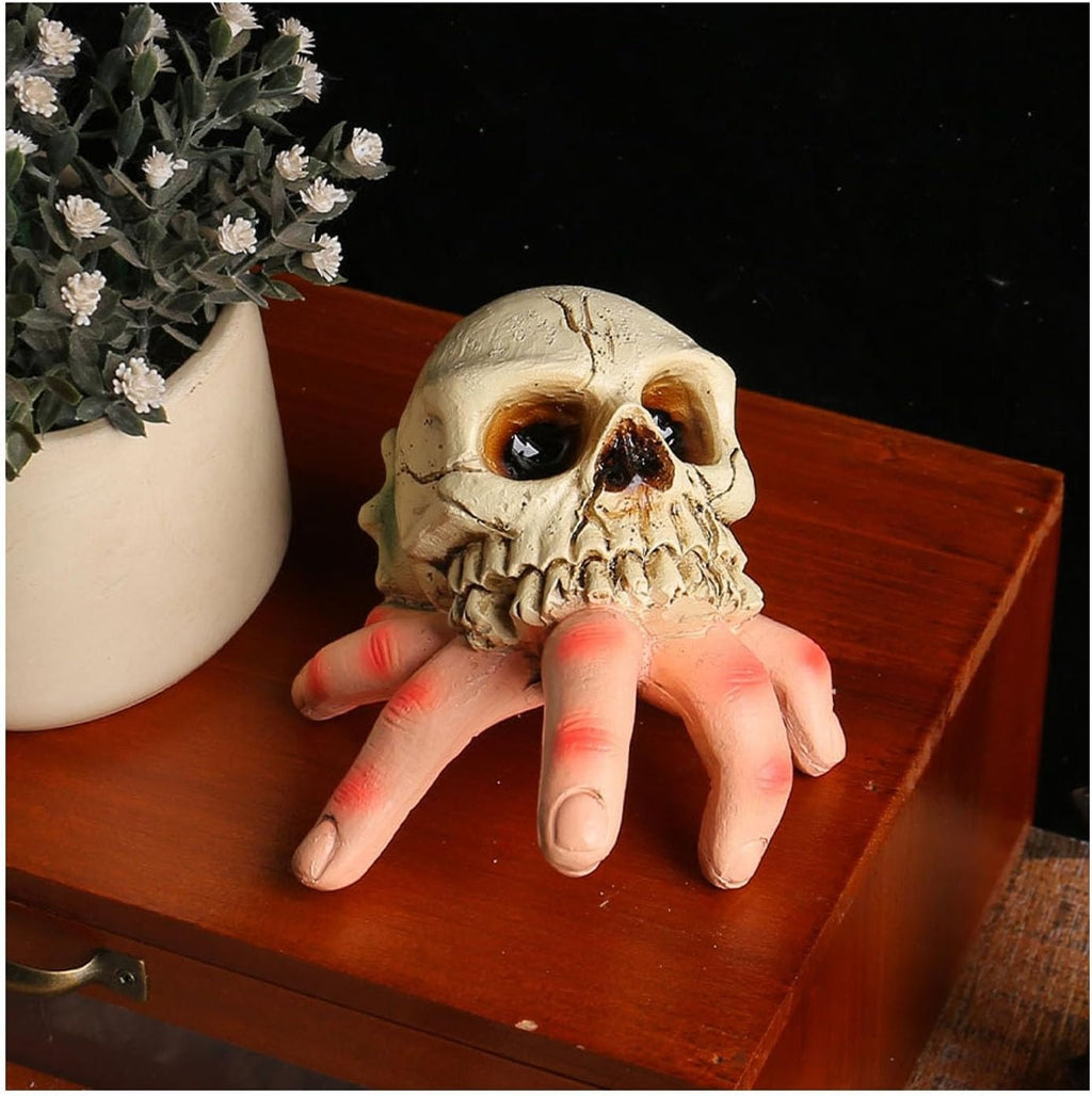 A Halloween prop of a skull with a creepy hand emerging from the mouth, resting on a wooden surface next to a potted plant.