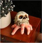 A Halloween prop of a skull with a creepy hand emerging from the mouth, resting on a wooden surface next to a potted plant.