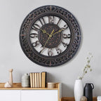 Decorative wall clock with intricate design on a gray wall, with books and vases on a shelf below.