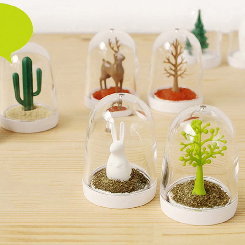 Five glass dome miniatures with rabbit, cactus, deer, and tree figurines on a wooden table, with a green speech bubble and small decor in the background.