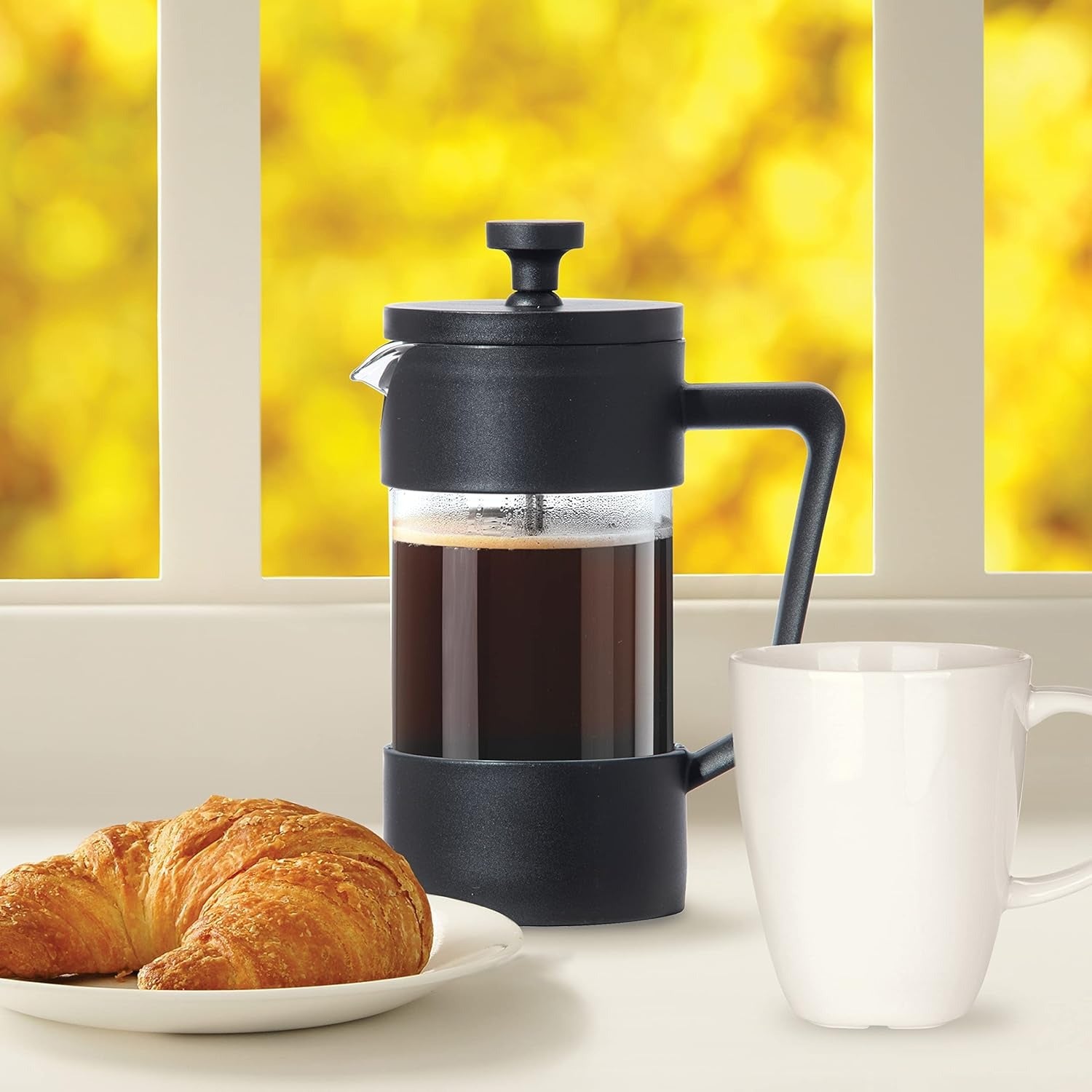 French press with coffee and a croissant on a table with a yellow window background
