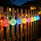 Multicolor solar-powered outdoor string lights hanging in front of a wooden picket fence at dusk, with glowing red, green, blue, yellow, pink, and light blue spherical bulbs.