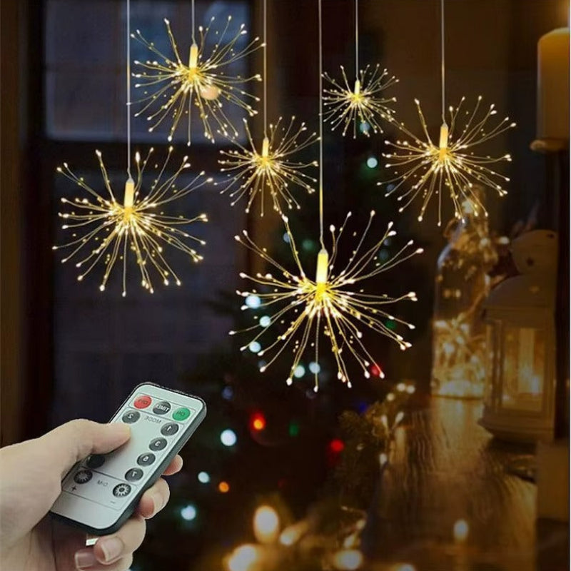 A set of hanging firework-shaped LED string lights glowing with warm white light, shown with a hand holding a remote control in a cozy indoor setting.