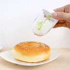 A hand holds a clear plastic sprinkler above a sesame-topped bun on a white plate, with white sprinkles falling onto the bun against a clean white background.