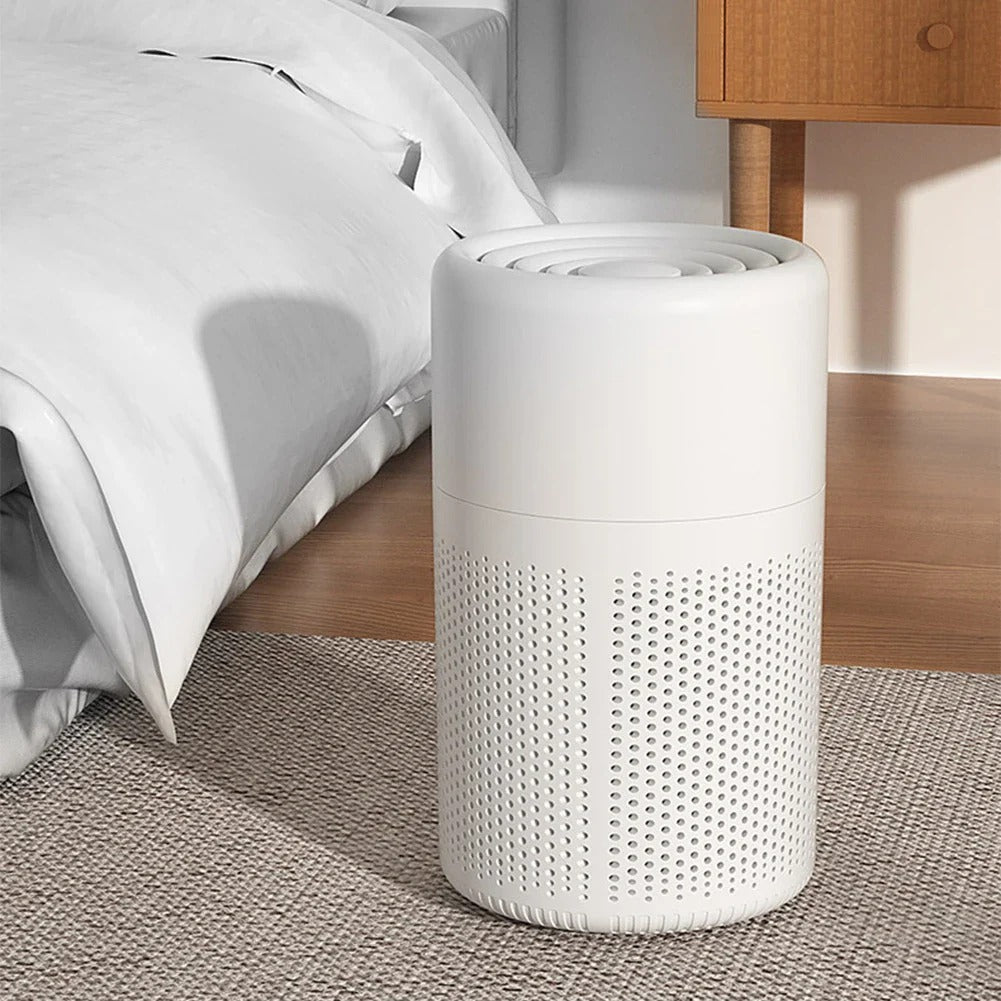 White cylindrical air purifier on a carpeted floor next to a bed in a bedroom.