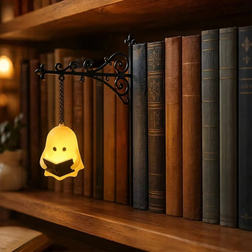 A small ghost-shaped hanging lamp glowing warm yellow, holding an open book, mounted on a black decorative bracket attached to a wooden bookshelf beside a row of antique-style books.