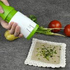 A hand holds a green and white manual vegetable chopper over a gray surface, with chopped green herbs falling onto a small cloth square. Two red tomatoes and a yellow potato sit behind the chopper, and the vegetable roots are still attached.