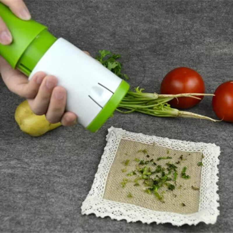 A hand holds a green and white manual vegetable chopper over a gray surface, with chopped green herbs falling onto a small cloth square. Two red tomatoes and a yellow potato sit behind the chopper, and the vegetable roots are still attached.