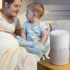 Woman and baby sitting on a couch with an air purifier on a table next to them.