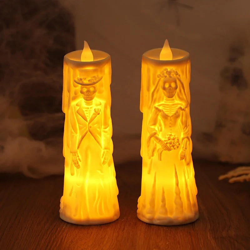 Two Halloween-style LED pillar candles shaped like skeleton bride and groom figures, glowing with warm yellow light on a wooden surface against a dark, spooky background.
