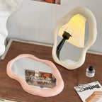 Two unique-shaped mirrors on a wooden surface with a lamp and magazine.