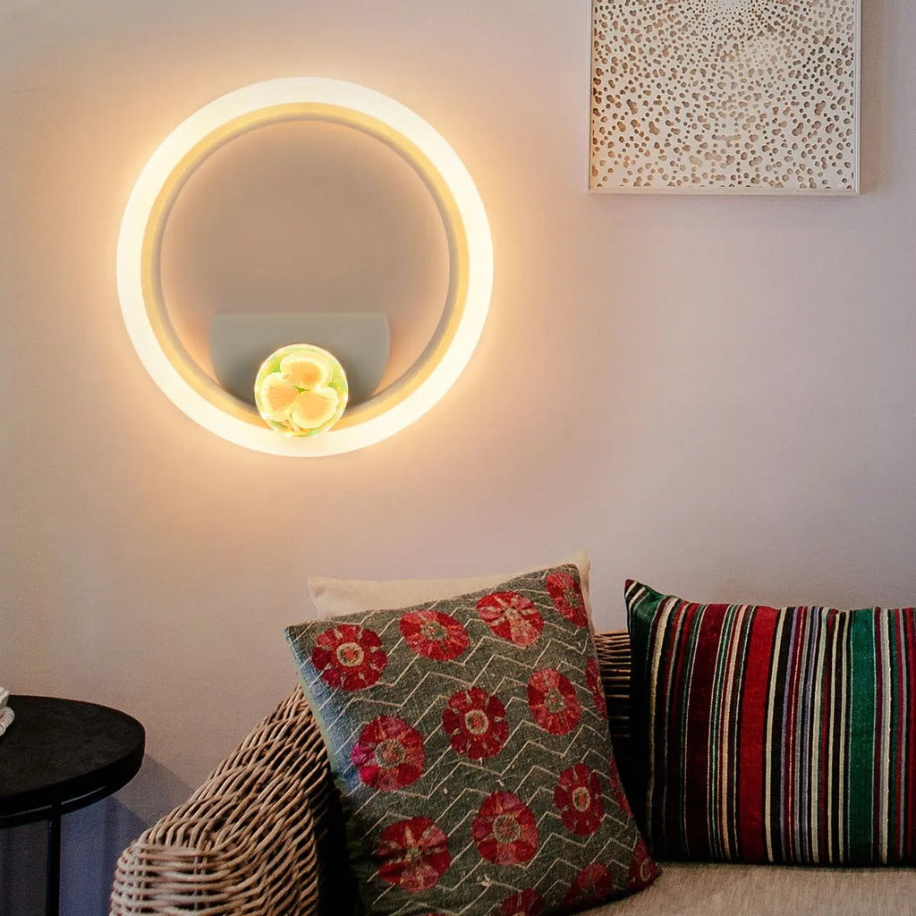 A modern circular LED wall light with a glowing warm white ring, mounted on a beige wall above a wicker sofa with patterned pillows.