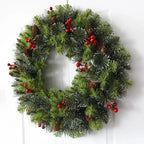 A festive artificial Christmas wreath with green pine needles, white snow dusting, brown pinecones, and clusters of red berries, hanging against a white paneled door.