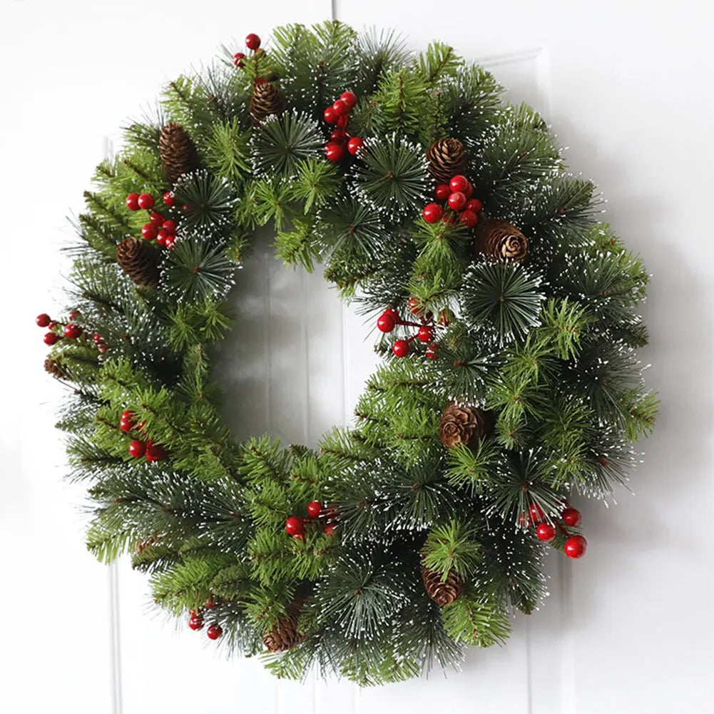 A festive artificial Christmas wreath with green pine needles, white snow dusting, brown pinecones, and clusters of red berries, hanging against a white paneled door.
