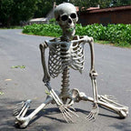 A life-size kneeling plastic skeleton decoration sitting on an asphalt road outdoors with greenery and buildings in the background.