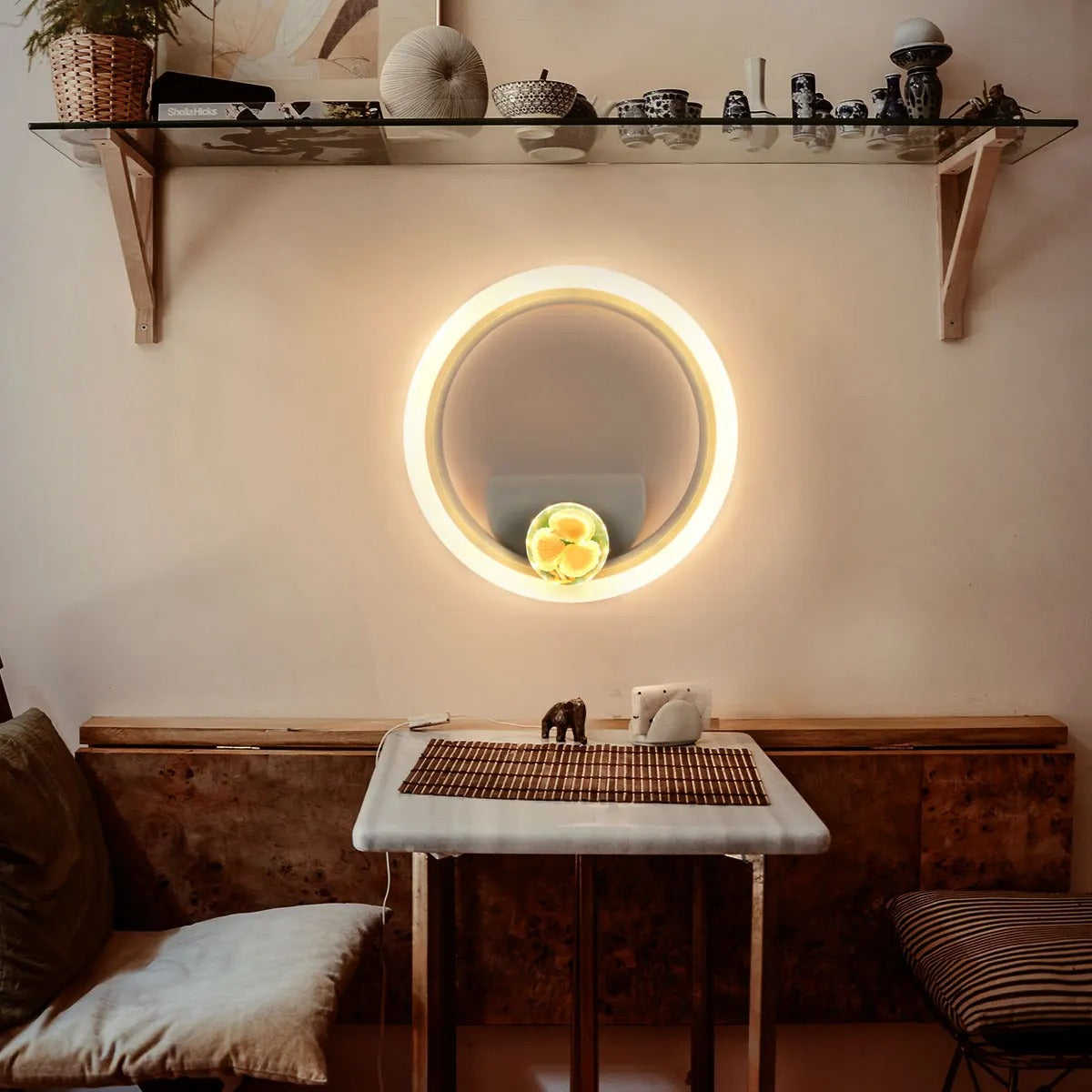 A glowing round wall mirror mounted on a beige wall above a small table in a styled interior. The mirror is illuminated with a warm light, and the room includes a glass shelf with decor, cushions, and a wooden bench.