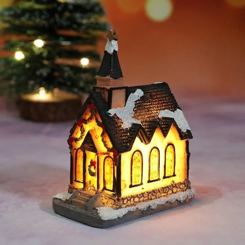 Decorative LED house with a warm glow, placed on a snowy surface with a blurred Christmas tree in the background.