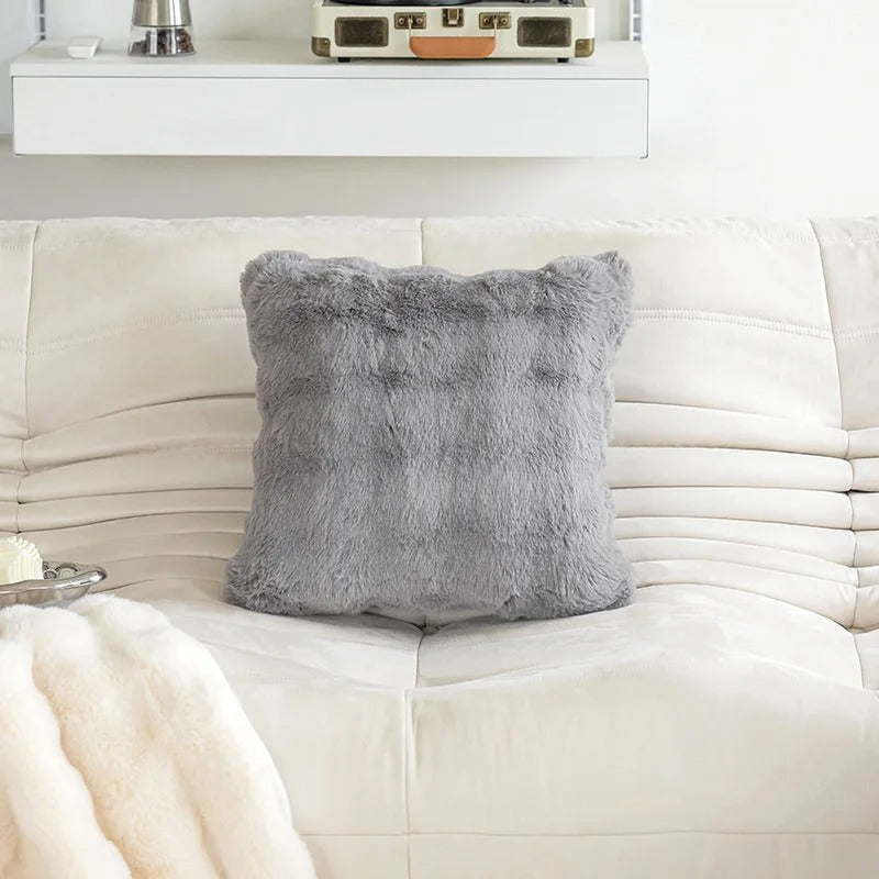 A square gray faux fur throw pillow resting on a cream-colored sofa in a modern living room setting.