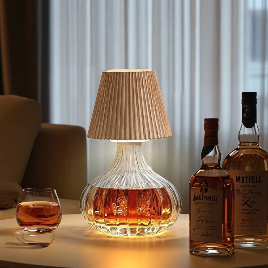 Glass lamp filled with a amber liquid, surrounded by bottles and a glass on a table.