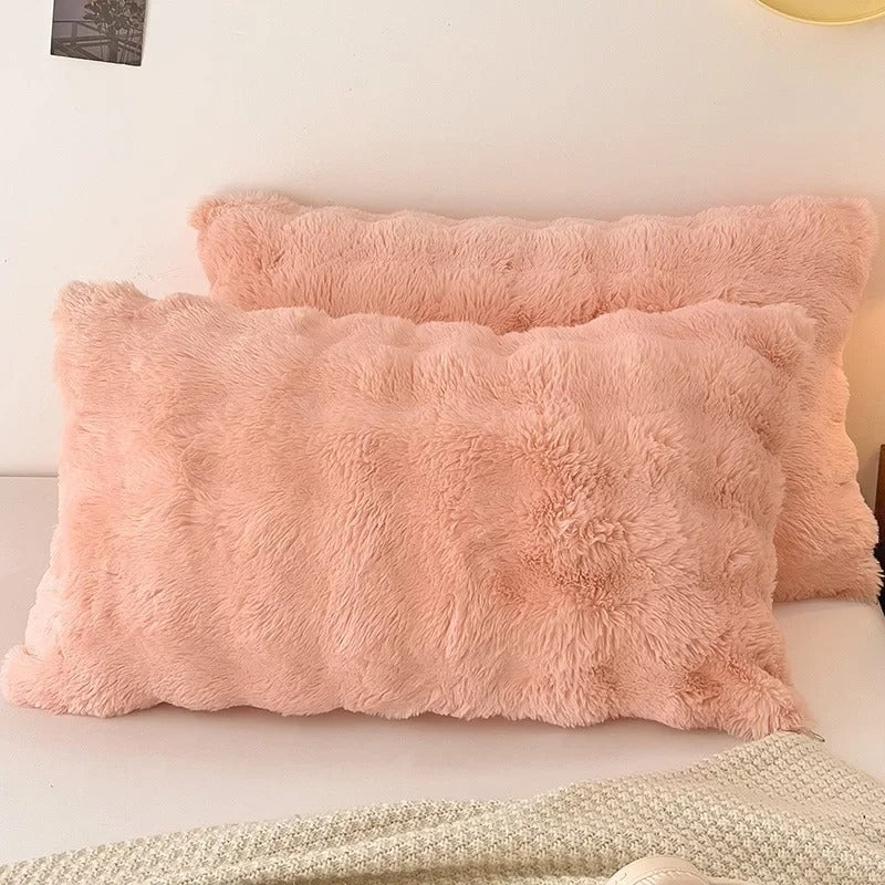 Two fluffy blush pink faux-fur pillows resting on a bed with a light-colored blanket and a soft wall background.